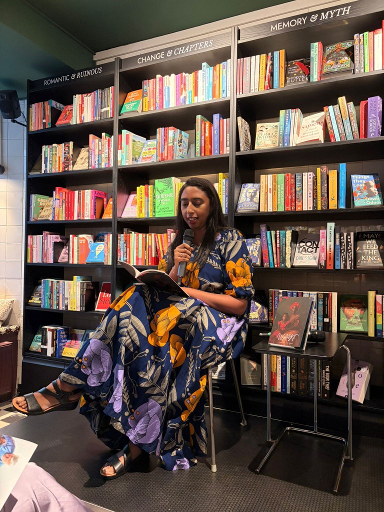 Sahra reading from a book at a microphone, with WAYF on the table beside her